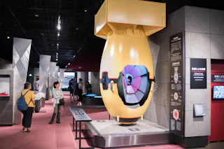 A large yellow bomb replica is displayed in a museum exhibit. Several visitors observe the exhibit, which features informational panels and modern lighting in a spacious room.