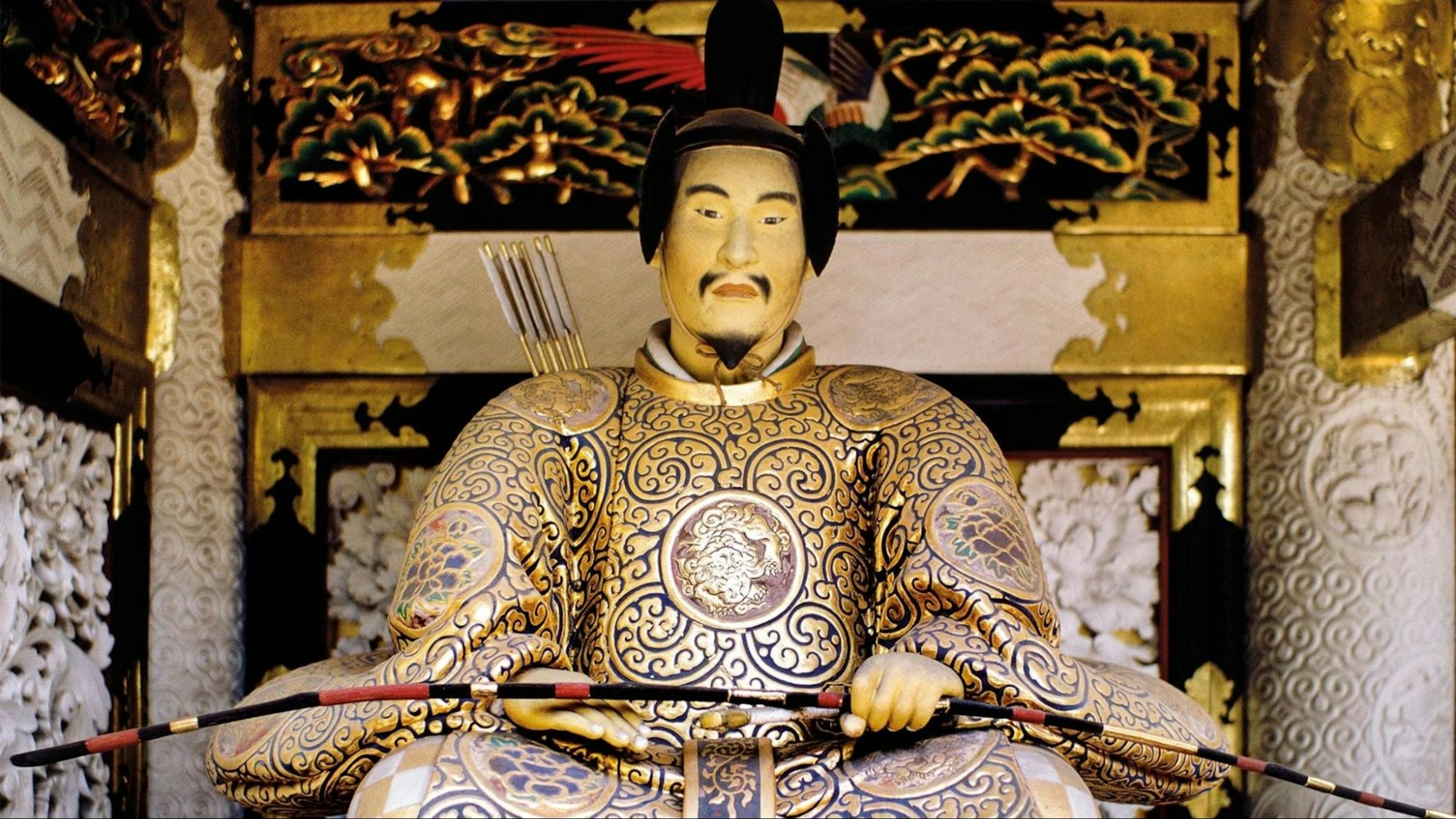 A regal golden statue of an ancient Japanese ruler seated on a throne, adorned in gleaming armor, with a crossbow and arrows displayed behind.