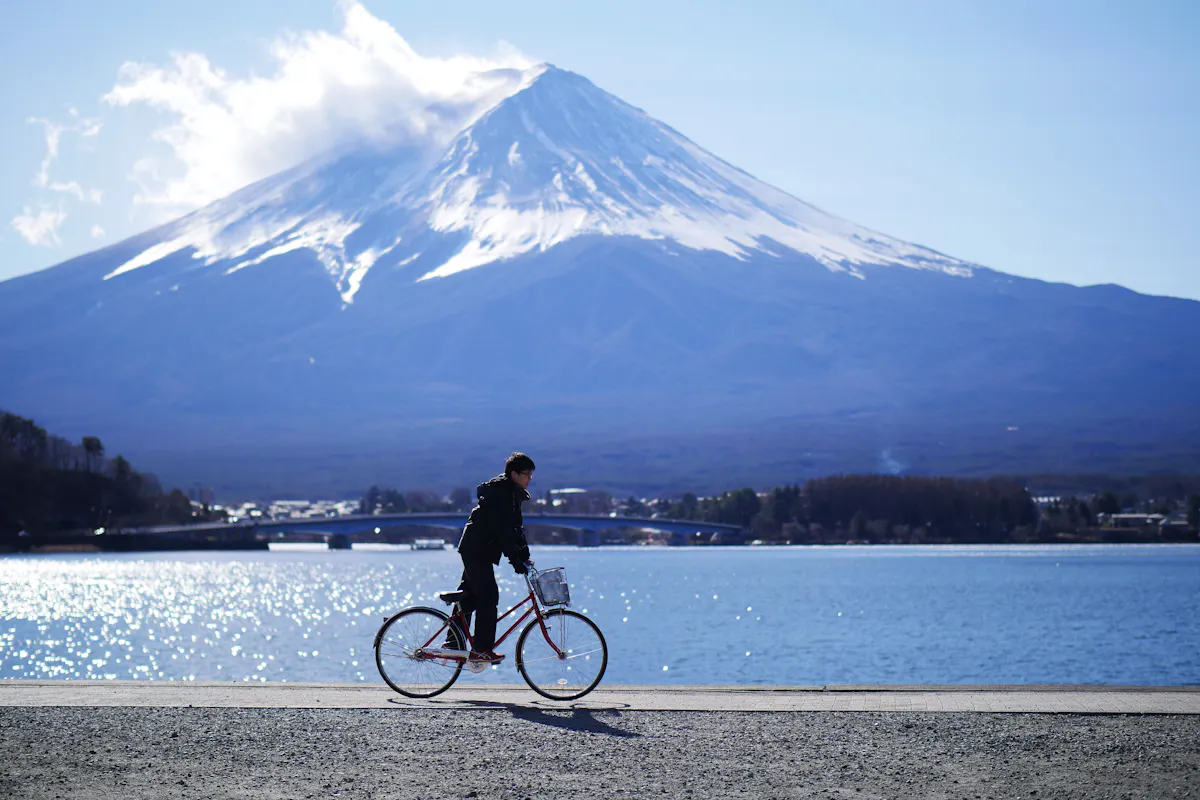 Mount Fuji for Adventure Seekers A person rides a bicycle along a path by a large body of water with Mount Fuji in the background. The sky is mostly clear with a few clouds near the mountain's peak. The scene is bright and serene, capturing a moment of peacefulness.