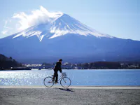 A person rides a bicycle along a path by a large body of water with Mount Fuji in the background. The sky is mostly clear with a few clouds near the mountain's peak. The scene is bright and serene, capturing a moment of peacefulness.
