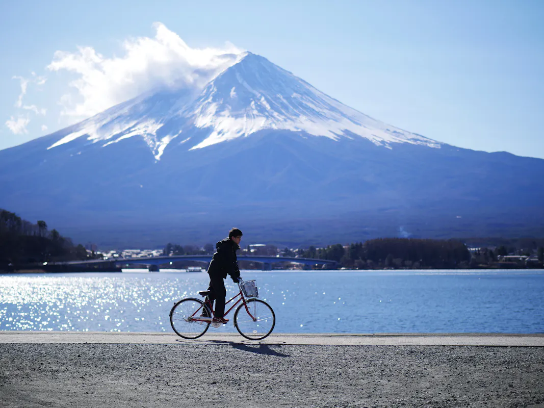 A person rides a bicycle along a path by a large body of water with Mount Fuji in the background. The sky is mostly clear with a few clouds near the mountain's peak. The scene is bright and serene, capturing a moment of peacefulness.