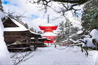 A snow-covered Buddhist temple complex, featuring a prominent red pagoda with an ornate spire, set against a backdrop of pine trees and a blue sky. The pathway is blanketed with snow, and traditional wooden buildings frame the serene winter scene.