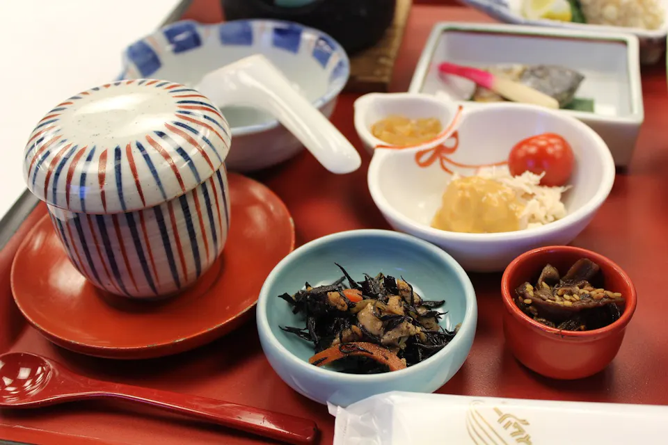 Shojin Ryori 색색의 일본식 식사가 쟁반 위에 놓여 있으며, 뚜껑이 있는 도자기 그릇, 빨간 숟가락, 해조류가 담긴 작은 그릇, 슬라이스된 채소가 담긴 접시, 둥글고 맛있는 초밥 롤, cherry 토마토, 소스가 담긴 작은 접시가 포함되어 있습니다. 주요 항목과 함께 다양한 작은 접시가 제공됩니다.