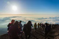 A large group of hikers in colorful gear ascend a steep, rocky mountain trail under a bright sun. A vast sea of clouds stretches out below them, with mountain peaks visible in the distance, creating a breathtaking, scenic vista.