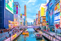 Colorful cityscape of Dotonbori in Osaka, Japan, with bright neon billboards, crowds on walkways, and boats carrying people along the canal under a blue sky.