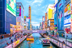Colorful cityscape of Dotonbori in Osaka, Japan, with bright neon billboards, crowds on walkways, and boats carrying people along the canal under a blue sky.