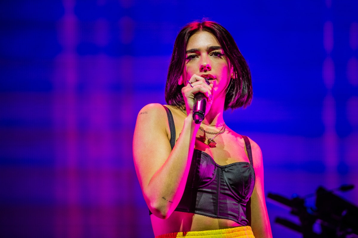 Dua Lipa A woman with short dark hair, wearing a black cropped top and yellow pants, is singing into a microphone on a brightly lit stage. The background features a mix of purple and blue hues. She is looking ahead with a focused expression.