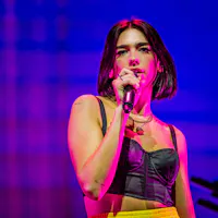 Dua Lipa A woman with short dark hair, wearing a black cropped top and yellow pants, is singing into a microphone on a brightly lit stage. The background features a mix of purple and blue hues. She is looking ahead with a focused expression.