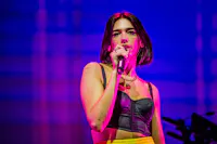 A woman with short dark hair, wearing a black cropped top and yellow pants, is singing into a microphone on a brightly lit stage. The background features a mix of purple and blue hues. She is looking ahead with a focused expression.