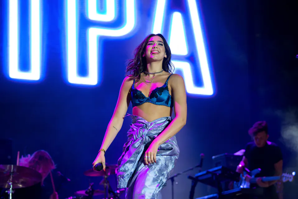 A performer with long dark hair, wearing a blue and black top and silver pants, smiles while holding a microphone on stage. In the background, vibrant blue and purple lights illuminate musical instruments and the letters "PA" are prominently displayed.