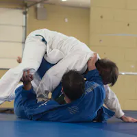 Groundwork practice on the mat, a regular part of judo training. Two judoka in white and blue gi grapple on a blue mat, one pinning the other from above in a bright training hall