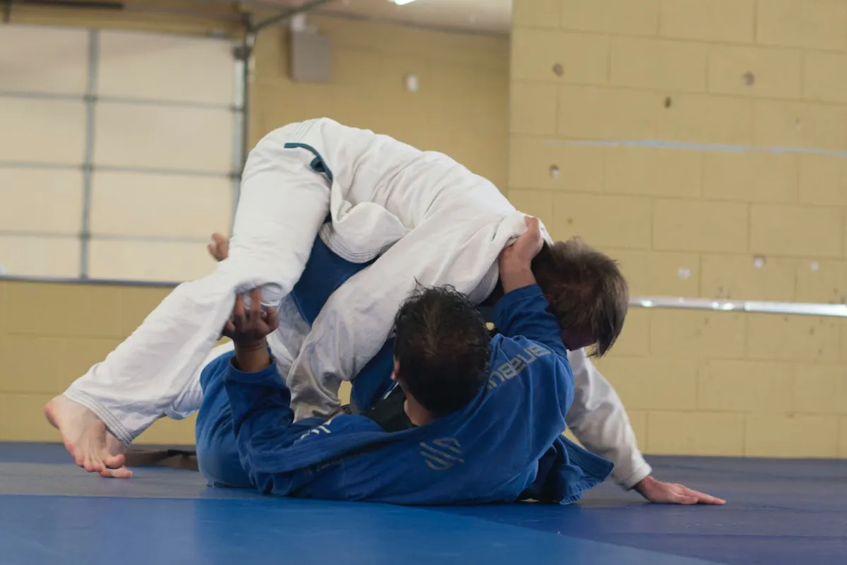 Groundwork practice on the mat, a regular part of judo training. Two judoka in white and blue gi grapple on a blue mat, one pinning the other from above in a bright training hall
