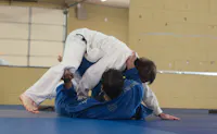 Two judoka in white and blue gi grapple on a blue mat, one pinning the other from above in a bright training hall