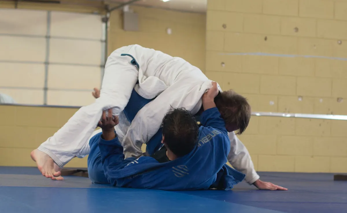 Two judoka in white and blue gi grapple on a blue mat, one pinning the other from above in a bright training hall