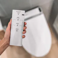 Smart Japanese Automated Toilet A person is holding a white remote control for a bidet toilet, showing options for rear, front, pulse, oscillating water streams, and a dryer function, along with stop and power buttons. The toilet is visible in the background.