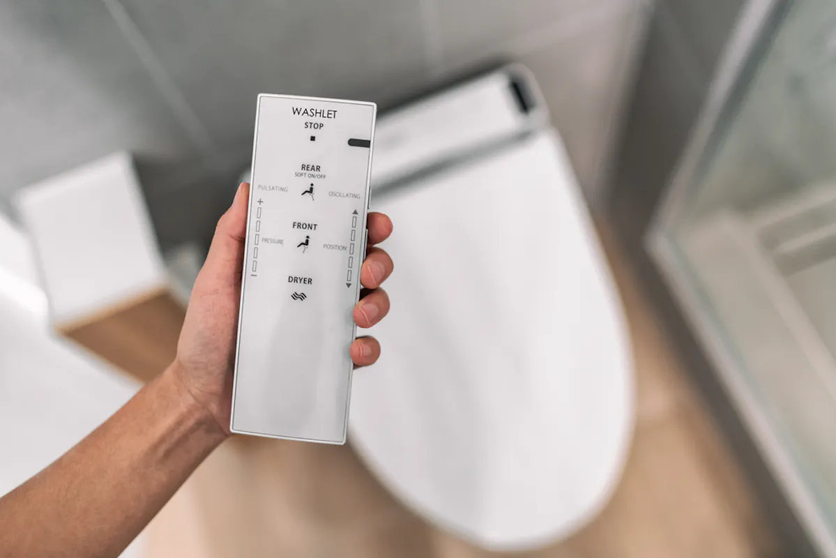A person is holding a white remote control for a bidet toilet, showing options for rear, front, pulse, oscillating water streams, and a dryer function, along with stop and power buttons. The toilet is visible in the background.