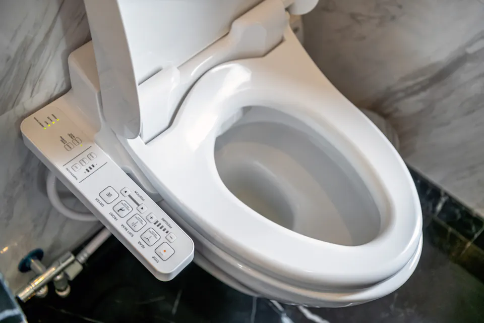 A modern toilet with a built-in bidet is shown. The bidet control panel on the left features multiple buttons and settings for various functions such as water pressure, nozzle positioning, and temperature adjustment. The bathroom has a marble wall and dark flooring.