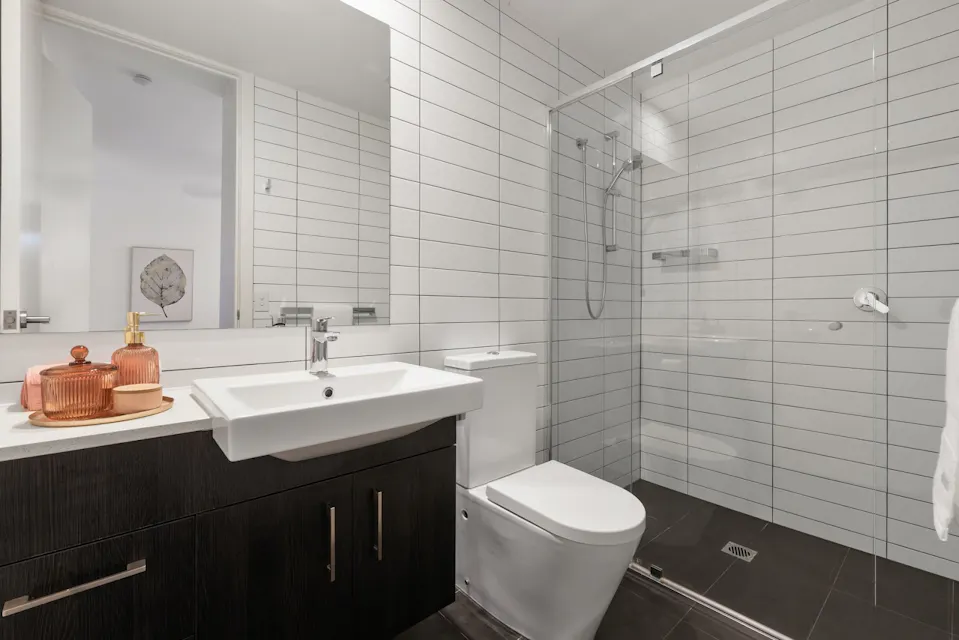 A modern bathroom featuring a white sink with a sleek faucet, a toilet, and a large glass-enclosed shower. The walls are tiled in white with dark grout. Below the sink is a dark wood cabinet. A white towel hangs on a hook, and decorative items sit on the counter.
