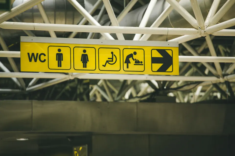 A yellow sign hanging from a ceiling with white structural beams indicates restrooms and related facilities. Icons show symbols for male, female, accessible, baby changing station, and an arrow pointing to the right.
