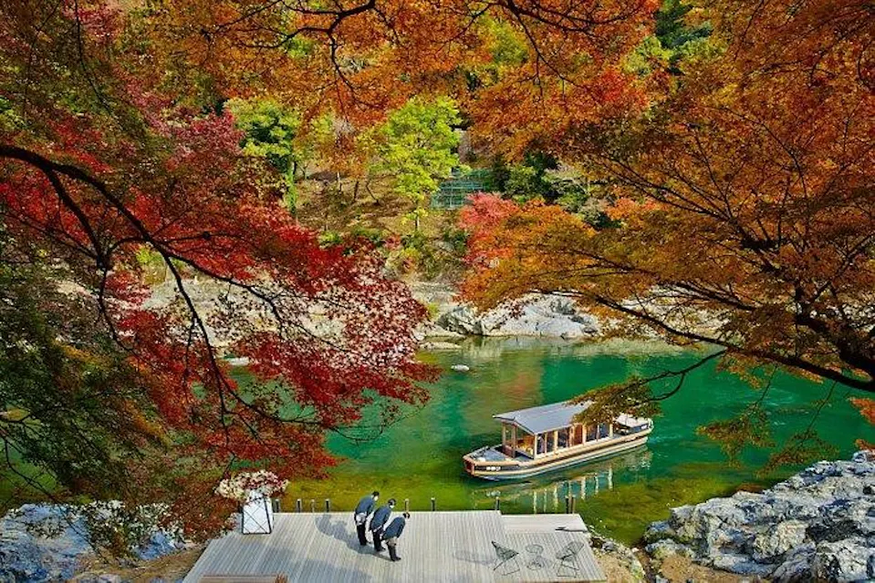 Hoshinoya Kyoto A serene autumn scene shows a small group of people on a wooden deck overlooking a clear, green river. A boat is docked by the deck, surrounded by vibrant red, orange, and yellow foliage. Large rocks line the riverbank, enhancing the natural beauty of the landscape.