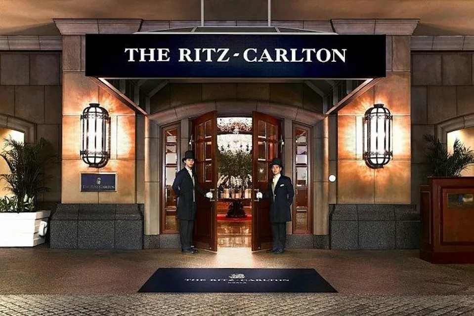 Ritz-Carlton, Osaka Two doormen in black coats stand on either side of the open entrance to The Ritz-Carlton, a luxury hotel. The entrance is warmly lit with elegant lighting, showing a glimpse of the lobby inside. The hotel's name is prominently displayed above the doors.