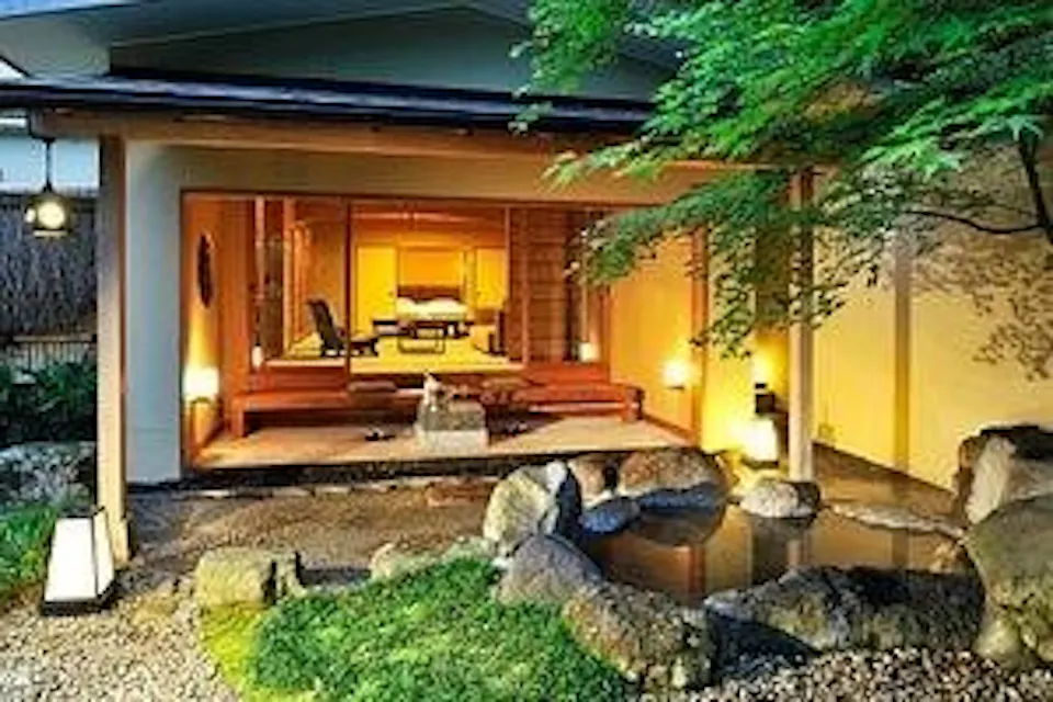 Gora Kadan, Hakone A cozy Japanese-style house with open sliding doors revealing a warmly lit interior. The house has a wooden deck overlooking a small garden with rocks and a tiny pond. Green foliage frames the scene, creating a serene and inviting atmosphere.