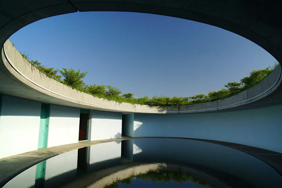 Benesse House, Naoshima A modern, circular building with a large open roof, allowing clear blue sky view. It has a shallow pool in the center, reflecting the surrounding structure, and plants growing along the top edge. The simplicity and open design create a serene and tranquil atmosphere.