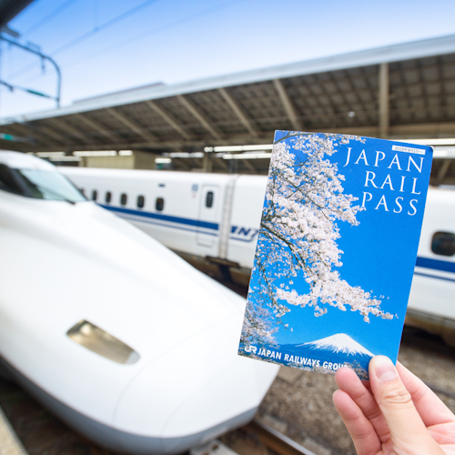 Japan Rail Pass A hand holds a Japan Rail Pass booklet with cherry blossoms on the cover in the foreground. In the background, a sleek white Shinkansen bullet train is stationed on the platform under a large, covered structure.