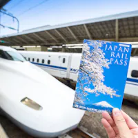 Japan Rail Pass A hand holds a Japan Rail Pass booklet with cherry blossoms on the cover in the foreground. In the background, a sleek white Shinkansen bullet train is stationed on the platform under a large, covered structure.