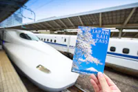 A hand holds a Japan Rail Pass booklet with cherry blossoms on the cover in the foreground. In the background, a sleek white Shinkansen bullet train is stationed on the platform under a large, covered structure.