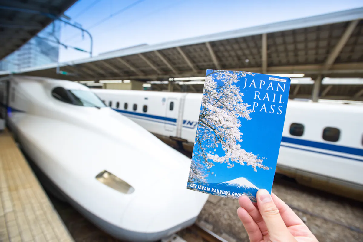 A hand holds a Japan Rail Pass booklet with cherry blossoms on the cover in the foreground. In the background, a sleek white Shinkansen bullet train is stationed on the platform under a large, covered structure.