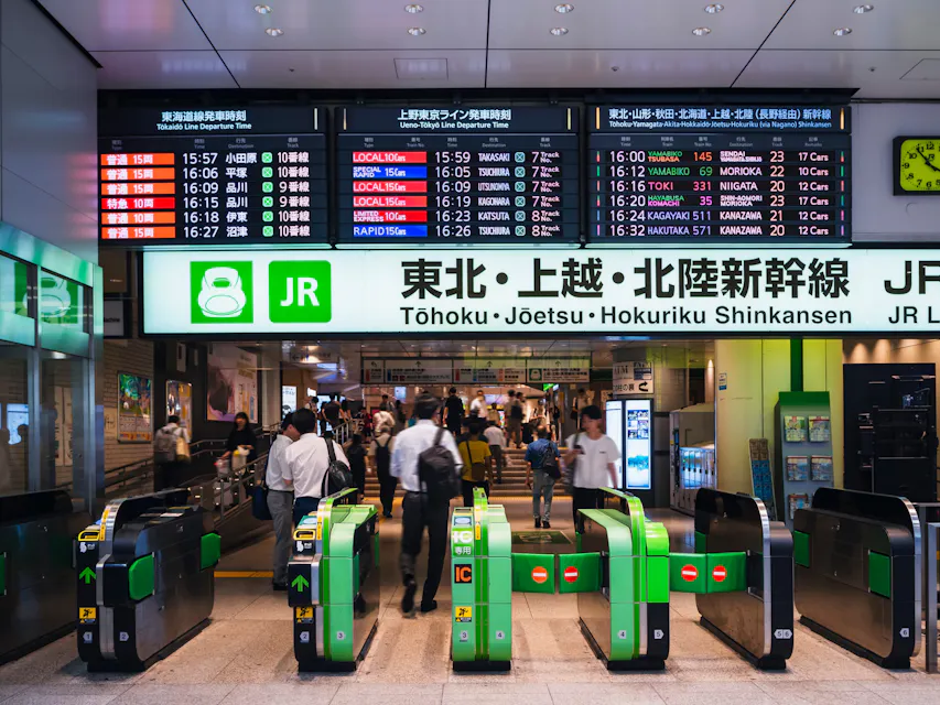 Crowded Japanese train station with green ticket gates in the foreground and several people passing through. Above, an electronic display shows train schedules in both Japanese and English for the Tohoku, Joetsu, and Hokuriku Shinkansen lines operated by JR East.