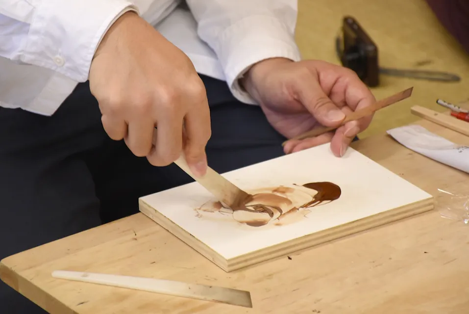A person in a white shirt is using a palette knife to mix paint on a wooden board. The workspace includes a wooden table, several tools, and materials, suggesting an environment set up for artistic activities or painting.