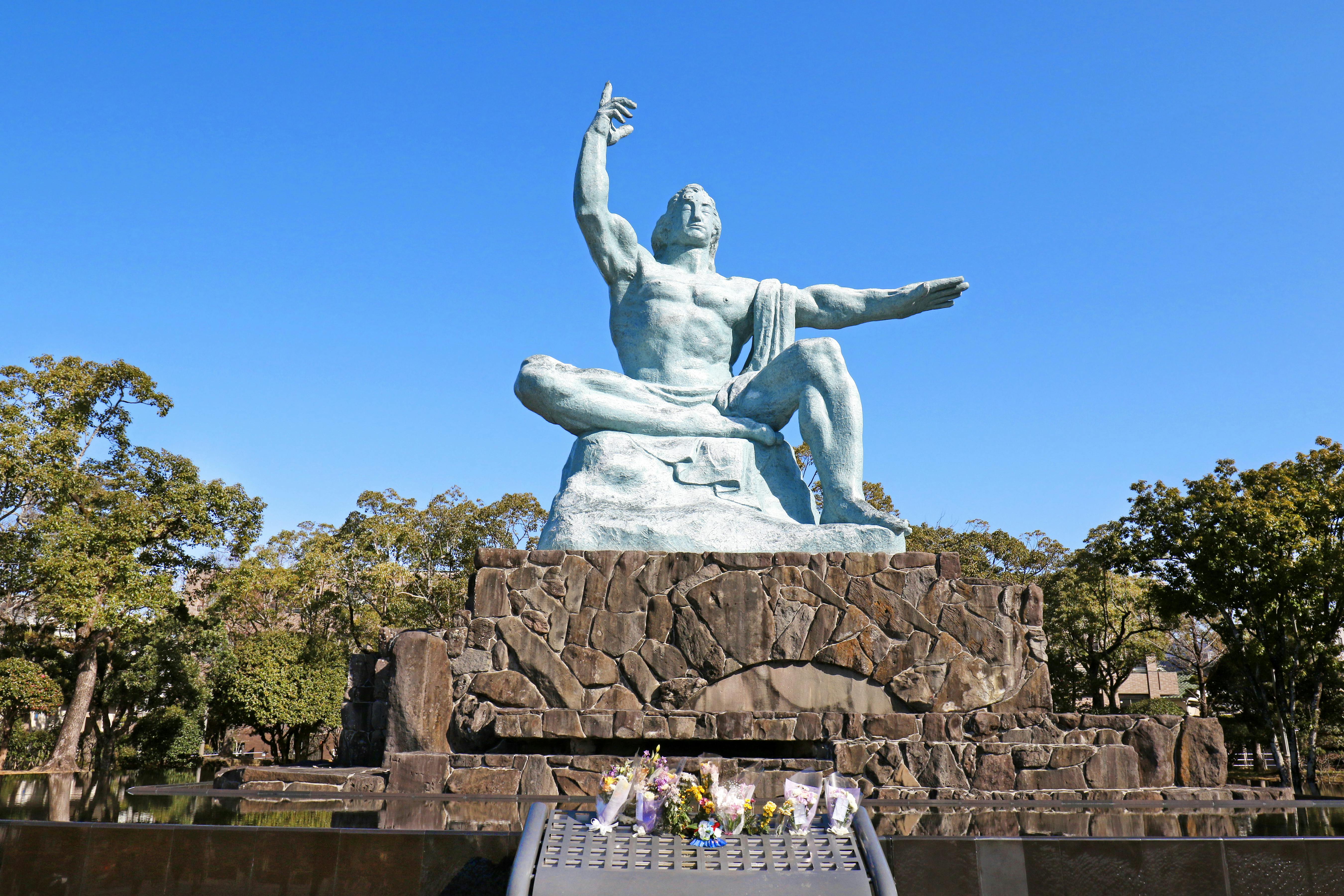 Large bronze statue of a seated man with one arm raised and the other extended, set on a stone pedestal with flowers in front, surrounded by trees and under a clear blue sky.