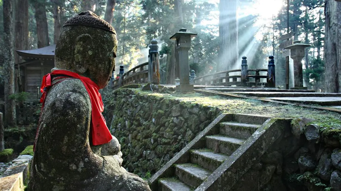 A serene stone Buddha statue with a red cloth around its neck sits in a tranquil forest setting. Sunlight filters through tall trees, illuminating moss-covered stone steps and a small bridge nearby. The peaceful atmosphere conveys calm and reflection.