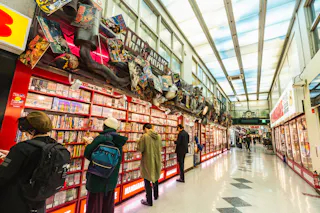 People browse manga bookshelves in a brightly lit Japanese store with colorful decorations and anime figures above the shelves. The floor is white with black accents and the atmosphere appears lively and busy.