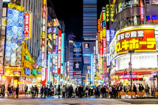 A busy street in Tokyo at night, filled with crowds and brightly lit with colorful neon signs and billboards. A large Godzilla head is visible atop a building in the background.