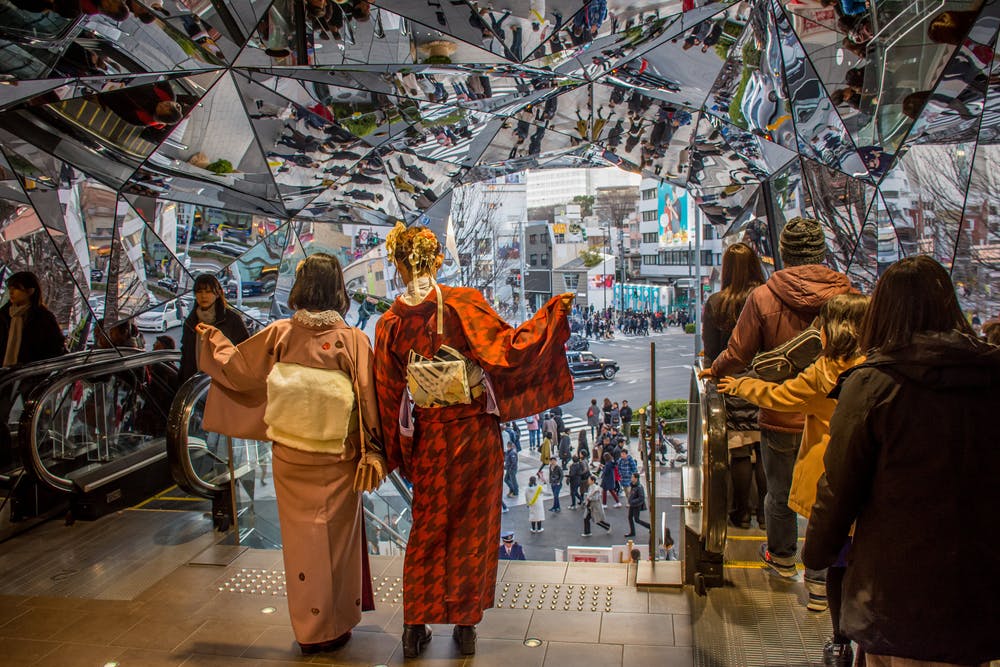 People in traditional Japanese clothing stand near escalators inside a building with mirrored walls and ceiling, reflecting the busy street scene and pedestrians outside. Crowds are visible both inside and outside.