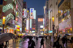 City street at night in the rain, crowded with people holding umbrellas. Tall buildings with bright, colorful signs and advertisements line both sides of the street, creating a lively urban atmosphere.