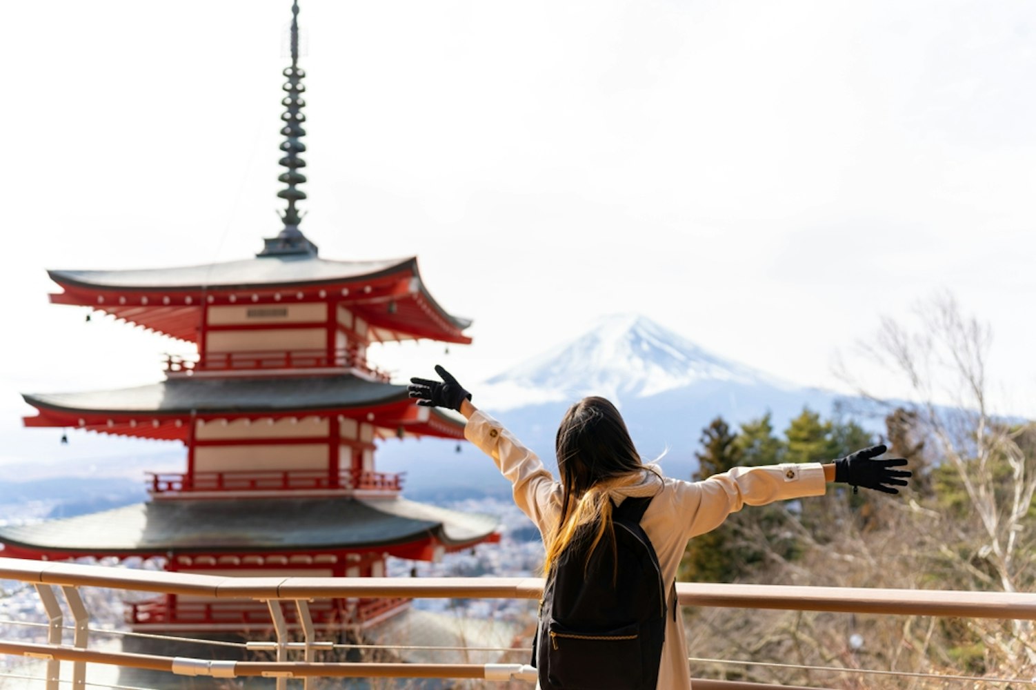A person wearing a black backpack and gloves stands with arms outstretched, facing a red pagoda. In the background, Mount Fuji is visible, partially covered with snow. The scene is bright and clear, with bare trees around.