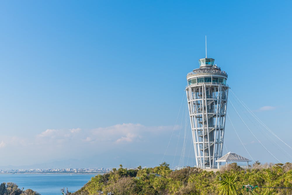 A tall, modern observation tower with a spiral structure stands on a green hill overlooking the sea under a clear blue sky.
