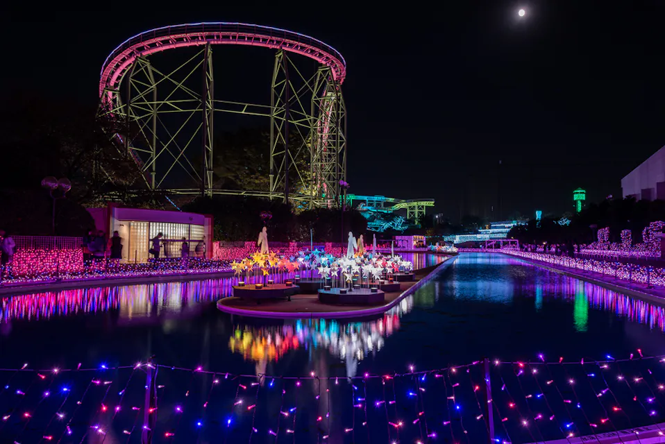 A vibrant nighttime scene of a theme park illuminated with colorful lights. A roller coaster is visible in the background, and a pool reflects multicolored lights. Decorative figures are arranged on platforms in the water.