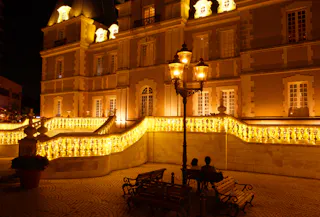 An illuminated building with ornate architecture at night, featuring glowing yellow lights along the stairs and railings. Two people sit on a bench in front of the building, under a vintage streetlamp.