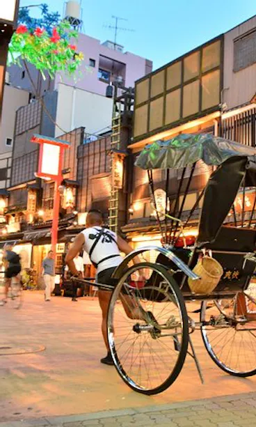 Asakusa Rickshaw Tour A traditional rickshaw is parked on a lantern-lit street lined with wooden Japanese buildings, featuring signs and warm glowing lights, creating an inviting evening atmosphere.