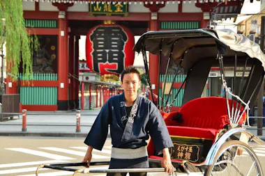 A rickshaw puller in traditional attire stands in front of a red temple gate with a large lantern at Senso-ji Temple in Asakusa, Tokyo, Japan.