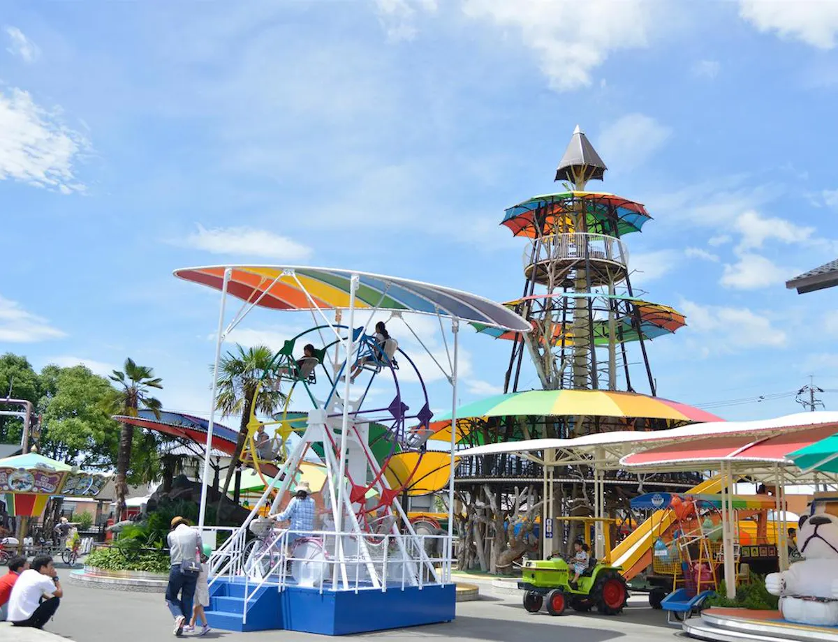 ACTLAND Creative Park A colorful amusement park with people enjoying rides under a bright blue sky. Visible attractions include a tall tower, a carousel, and various play structures with vibrant, multicolored canopies.