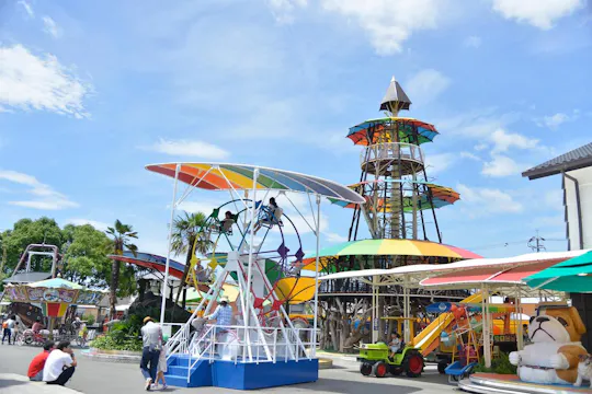A colorful amusement park with people enjoying rides under a bright blue sky. Visible attractions include a tall tower, a carousel, and various play structures with vibrant, multicolored canopies.