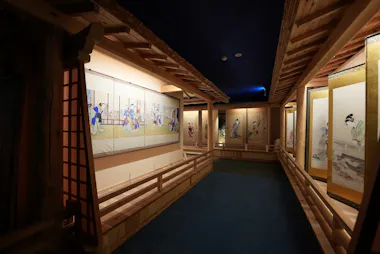 A dimly lit hallway in a museum displays traditional Japanese scroll paintings along wooden walls, with soft lighting highlighting the artwork and a blue carpeted floor below.