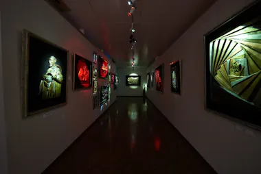A dimly lit art gallery hallway displays illuminated framed artworks on both walls, including abstract designs and a sculpture of a person, creating a dramatic, moody atmosphere.
