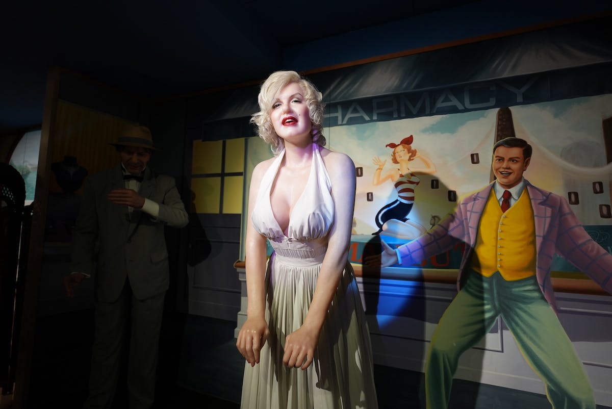 A wax figure of a woman in a white dress with blonde hair stands smiling, with two other figures and a colorful mural featuring the word "PHARMACY" in the background.